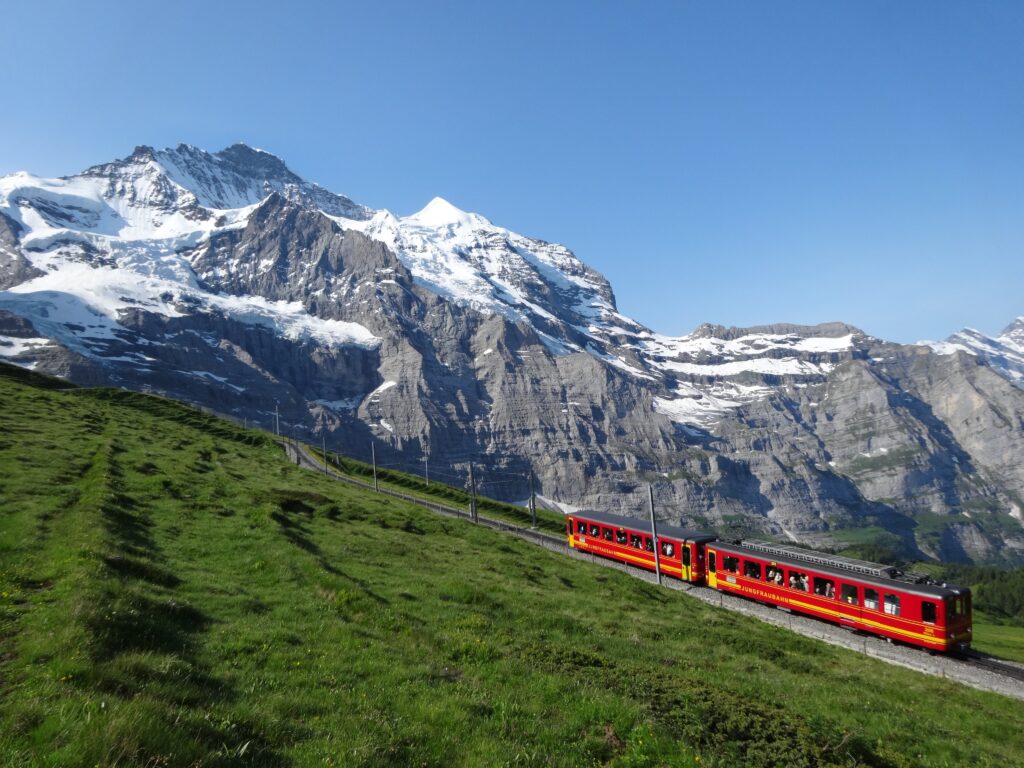 青い空に、白く冠雪したユングフラウと緑の草原の間を登る、赤い登山鉄道